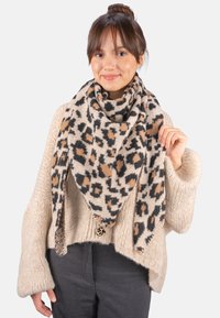 Écharpe en imprimé léopard dans des tons beige, noir et marron. Texture en tricot douce, forme triangulaire, drapée autour des épaules du modèle.