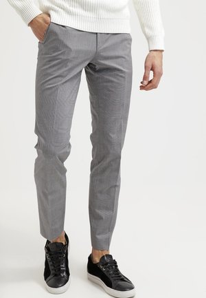 Mann trägt slim-fit graue karierte Hose, weißen Pullover und schwarze Low-Top-Sneaker, steht vor weißem Hintergrund.