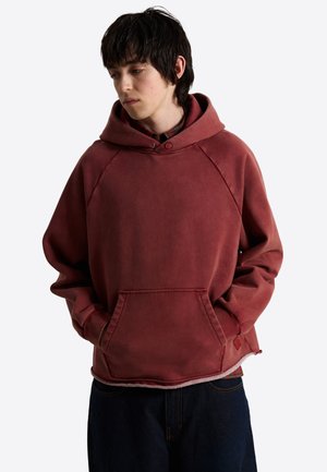 Junger Mann mit schwarzen Haaren, der ein ausgewaschenes rotes Kapuzensweatshirt mit Fronttasche und dunkle Hose trägt, schaut nach unten links.