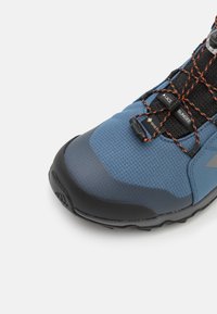 Botas de exterior azules con parte superior texturizada, puntera de goma negra y detalles en naranja en los cordones. Diseñadas para durabilidad y tracción.