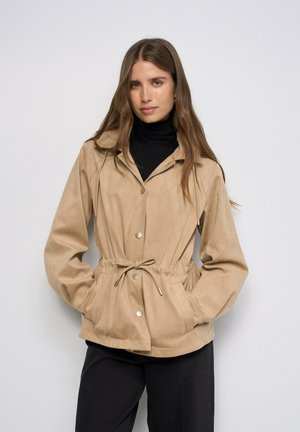 Mujer vistiendo una chaqueta beige abotonada con cintura ajustable sobre un jersey de cuello alto negro y pantalones negros, de pie frente a un fondo liso.