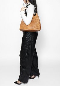 Geweven leren handtas in warm bruin met een gestructureerde vorm, voorzien van dubbele zwarte hengsels en een gestructureerd oppervlak.