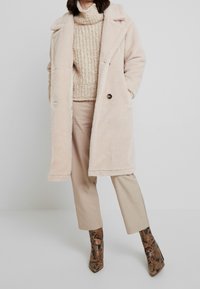 Beige fuskpälsjacka med notch-krage, dubbelknäppt design och svarta knappar, tillsammans med en texturerad krämfärgad tröja och bruna byxor.