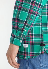 Camicia a quadri verde con motivi rosa, neri e bianchi, polsino abbottonato con etichetta. Realizzata in un tessuto testurizzato.