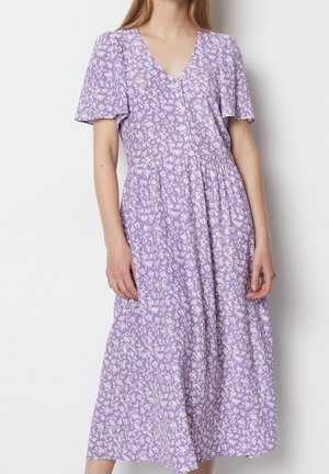 Femme portant une robe midi lavande à imprimé floral blanc, col en V, manches courtes volantes et taille froncée, debout devant un fond blanc.