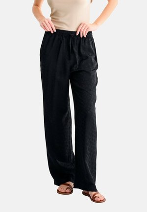 Femme portant un pantalon noir ample à motifs avec une taille élastique et un haut beige sans manches, debout avec les mains près de la taille.