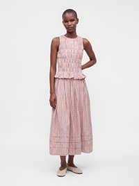 Woman wearing a sleeveless pink and white checkered dress with a ruffled waist and cream flat shoes standing against a plain background.