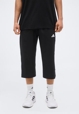 Person wearing black Adidas cropped pants, white Adidas socks, and white Adidas sneakers, standing on a plain white background.