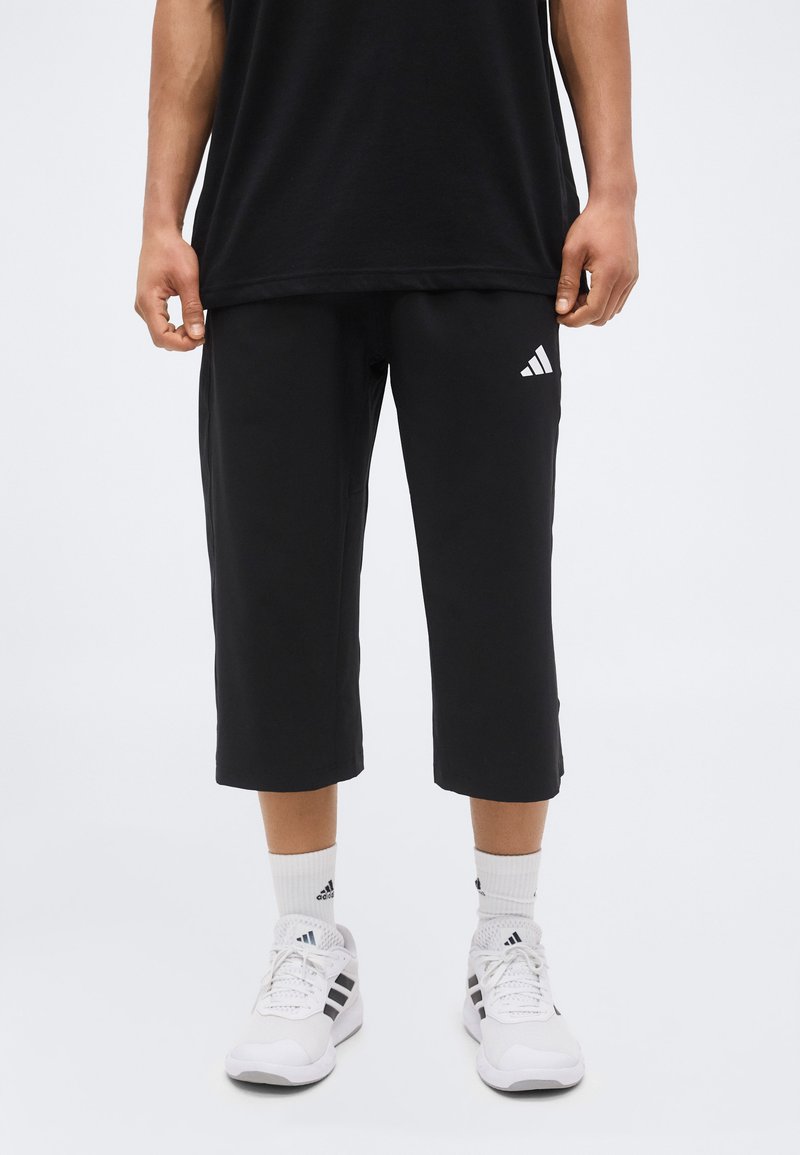 Personne portant un pantalon court noir Adidas, des chaussettes blanches Adidas et des baskets blanches Adidas, debout sur un fond blanc uni.