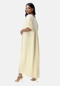 Robe longue et texturée de couleur jaune clair avec des manches courtes, un col rond et une coupe ample, associée à des talons beiges à lanières.