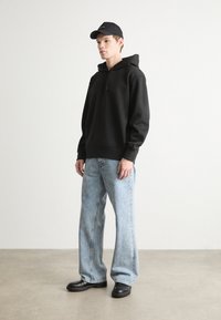 Hanorac negru cu glugă și buzunar tip kangaroo, asortat cu blugi din denim albastru deschis, cu croială largă și pantofi din piele negri. Look casual, modern.