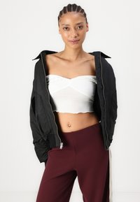 Veste bomber noire avec zip, portée ouverte sur un top court blanc avec un ourlet en volants, associée à un pantalon bordeaux ajusté.