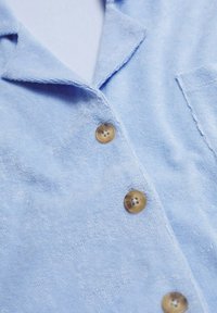 Světle modrá froté košile s límečkem, hnědými knoflíky vpředu a náprsní kapsou na pravé straně.