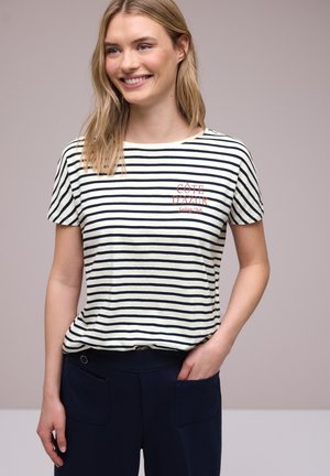 Lächelnde Frau, die ein weiß-navy gestreiftes T-Shirt mit dem Text „Côte D’Azur Sailing Club“ trägt, navy blaue Hose, steht mit einer Hand in der Tasche.