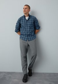 Blau-grün kariertes Hemd mit Knöpfen, graue Anzughose und schwarze Schuhe. Das Hemd hat hochgekrempelte Ärmel; glatter Stoff und taillierter Schnitt.