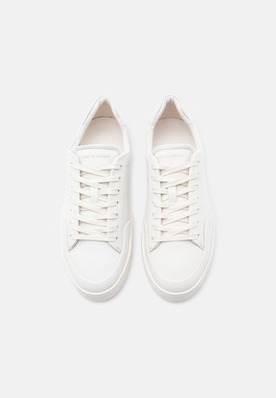 Λευκά αθλητικά παπούτσια με πάνω μέρος από λείο δέρμα, υφασμένους τόνους, στρογγυλεμένη μύτη και επίπεδα κορδόνια. Μινιμαλιστικός σχεδιασμός με διακριτικό λογότυπο στη γλώσσα.