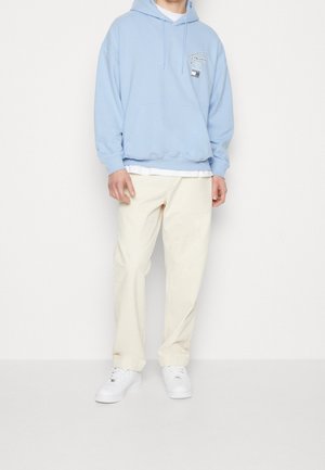 Felpa azzurro chiaro con cappuccio, tasca frontale e logo, abbinata a pantaloni color crema e scarpe da ginnastica bianche. Tessuto morbido e vestibilità comoda.