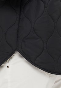 Zwarte gewatteerde vest met een golfpatroon, met een puntige onderzoom en zichtbare stikdetails, gedragen over een witte shirt.