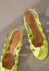 Keltaiset slingback-sandaalit, joissa on leikattu muotoilu, sileä kiiltävä pinta ja pehmustettu ruskea pohjallinen, jossa on kohokuvioitu brändäys.