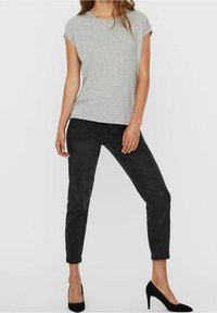 Donna che indossa una t-shirt grigio chiaro a maniche corte, jeans neri a vita alta slim e scarpe con tacco alto nere, in piedi con una gamba leggermente piegata.