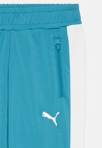 Turkosa sportbyxor med elastisk midja, sidofickor, vita sidodetaljer och en vit logotyp på vänster ben. Slät textur.