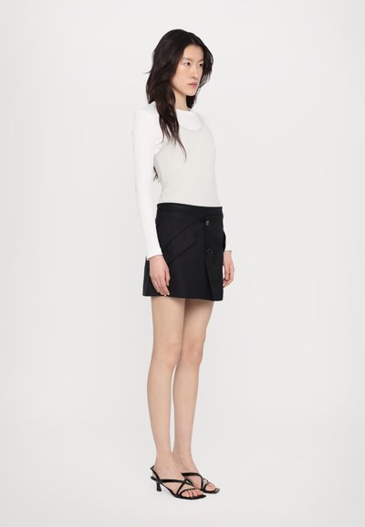 Melna īsa svārku ar pleciem, divi pogas, apvienota ar pieguļošu baltu garām piedurknēm topo. Modelis valkā melnas sandales ar siksniņām.