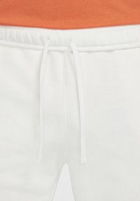 White sweatpants with a drawstring closure, smooth texture, and tapered legs. An orange shirt is partially visible above the waistband.