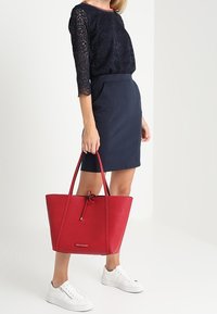 Borsa tote in pelle rossa con superficie texture, doppi manici e chiusura a cordoncino, abbinata a un top in pizzo blu navy e una gonna a tubino.