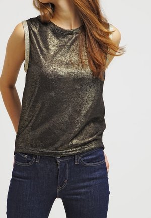 Kvinde iført en ærmeløs, metallisk gylden glimtende top kombineret med mørkeblå tætsiddende jeans, stående mod en ensfarvet baggrund.