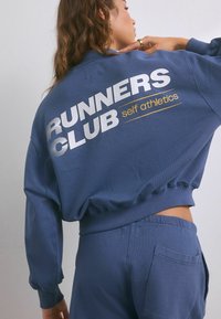 Felpa blu con polsini e orlo a coste, con la scritta "RUNNERS CLUB" in bold bianco e "self athletics" in oro. Materiale leggero, vestibilità comoda.