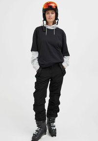 Black hoodie over a gray sweatshirt, black ski pants, and ski boots. The helmet has a reflective orange visor. All pieces are casual and functional.