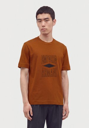 Hombre joven con una camiseta Emporio Armani de color óxido con texto del logo en negro, de pie contra un fondo liso de color gris claro.