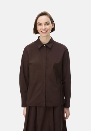 Donna sorridente con capelli castani corti, indossa una camicia marrone scuro con bottoni e una gonna coordinata su uno sfondo chiaro uniforme.
