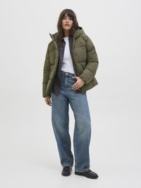 Doudoune olive avec capuche, associée à un t-shirt blanc et une chemise à carreaux en dessous. Jean bleu ample et chaussures noires.