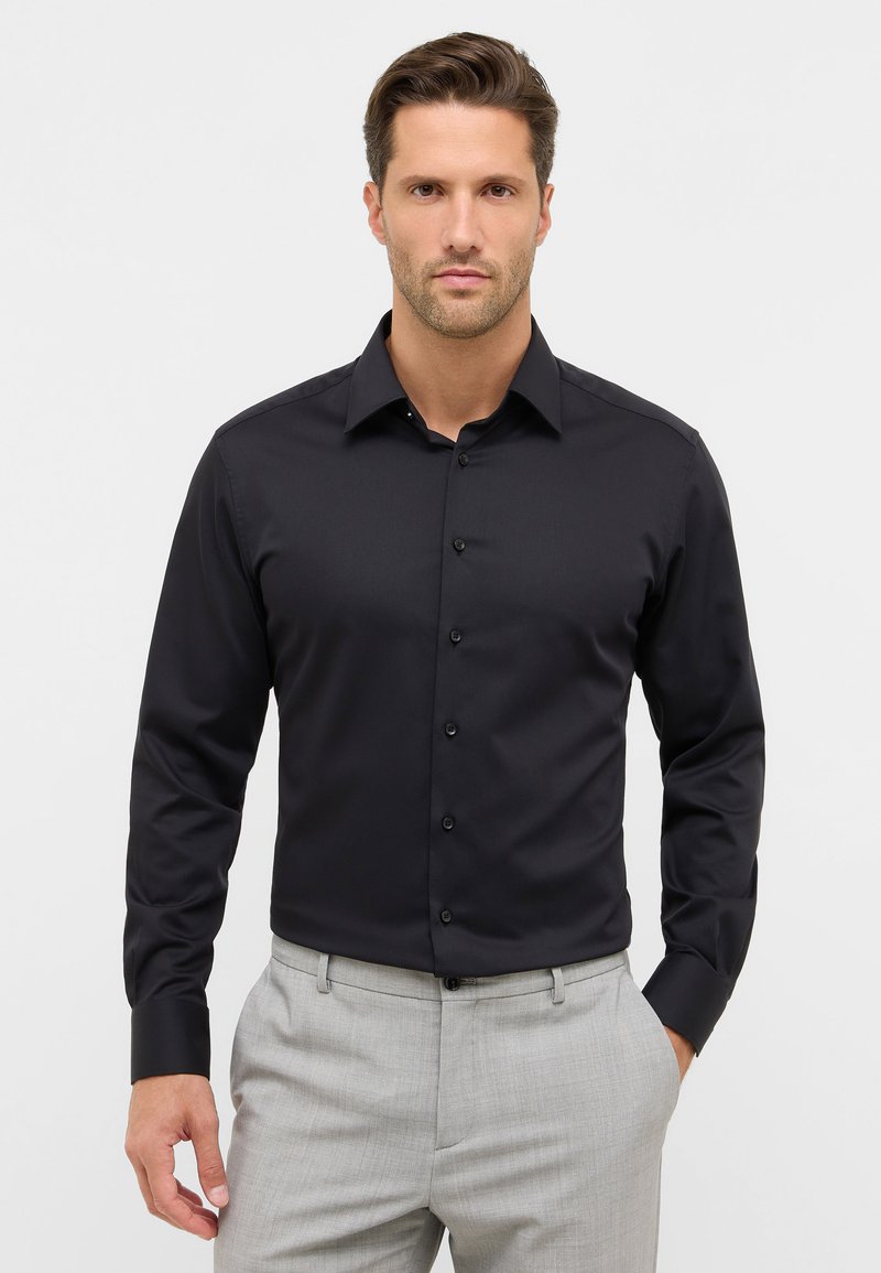 Camicia nera con colletto classico, texture liscia, maniche lunghe e polsini con linguetta, abbinata a pantaloni grigi chiari su misura.