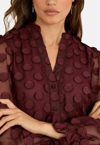 Kvinna iklädd en genomskinlig bourgogneblus med upphöjda prickar, volangärmar och en V-hals med tygklädda knappar, armarna i kors.