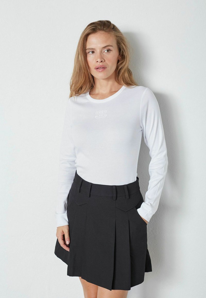 Top bianco a maniche lunghe con scollatura rotonda e logo ricamato, abbinato a una gonna nera plissettata con vita con cintura e tasche laterali.