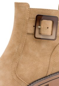 Beige Wildleder-Ankle-Boot mit einer quadratischen braunen Schnalle, detaillierter Steppnaht und einer strukturierten Sohle. Die Gesamtform ist schlank mit einer geschwungenen Silhouette.