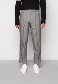 Harmaat, ohuet raidalliset housut, joissa on laskostettu etuosa, räätälöity istuvuus ja lyhyt pituus, yhdistettynä valkoisiin tennareihin ja mustaan neulepuseroon.