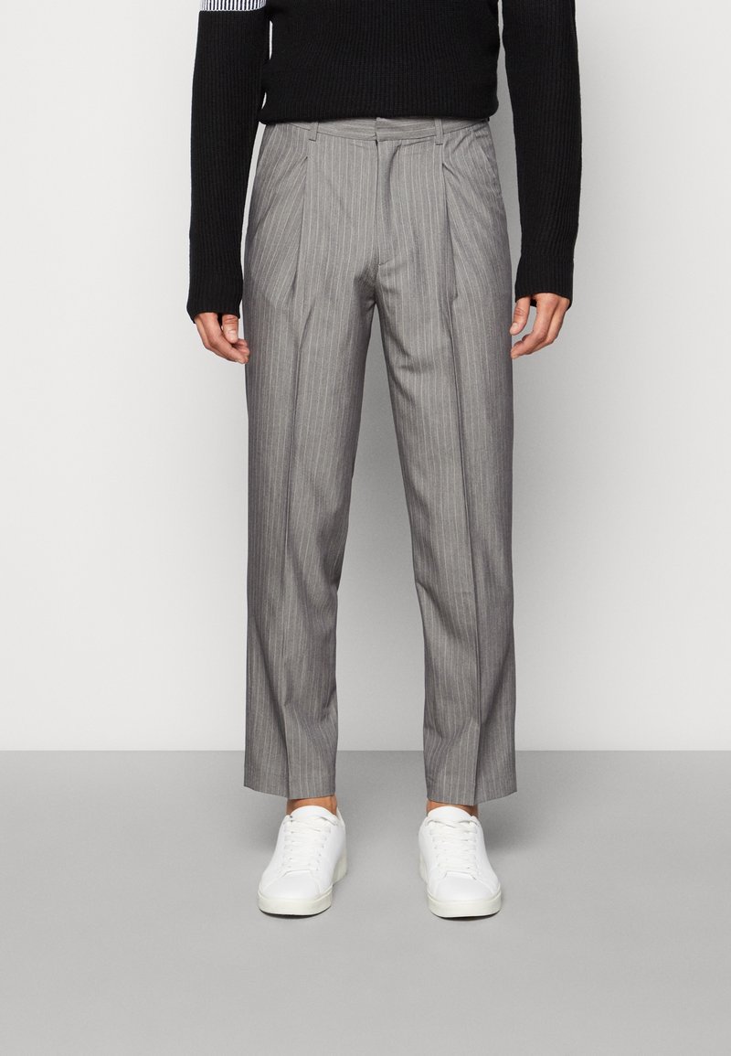 Harmaat, ohuet raidalliset housut, joissa on laskostettu etuosa, räätälöity istuvuus ja lyhyt pituus, yhdistettynä valkoisiin tennareihin ja mustaan neulepuseroon.