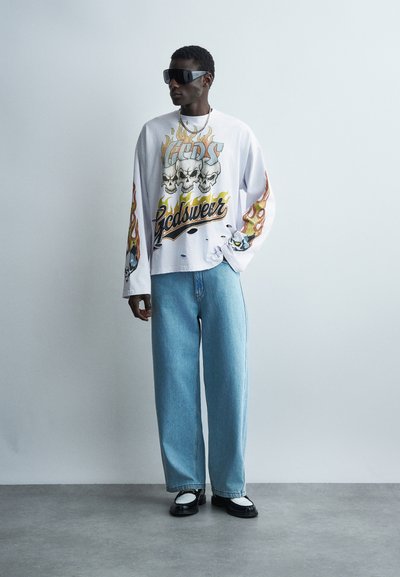 Άνδρας που φοράει υπερμεγέθη λευκό φούτερ με γραφικά κρανίου και φλόγας, ανοιχτό μπλε τζιν φαρδιάς γραμμής, ασπρόμαυρα παπούτσια και μεγάλα μαύρα γυαλιά ηλίου.