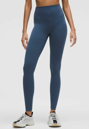 Hochtaillierte blaue Leggings aus einem glatten, dehnbaren Material, mit passgenauer Passform und nahtlosem Bund. Getragen mit weißen Sneakers.