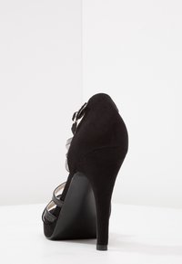 Černá semišová vysoká bota s podpatkem, která má více pásků s kovovými detaily a špičatý tvar špičky, umístěná na bílém pozadí.