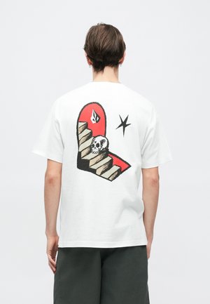 T-shirt en coton blanc présentant un graphique coloré d'un crâne sur des escaliers dans une arche rouge. Accents noirs et un petit détail de logo inclus.