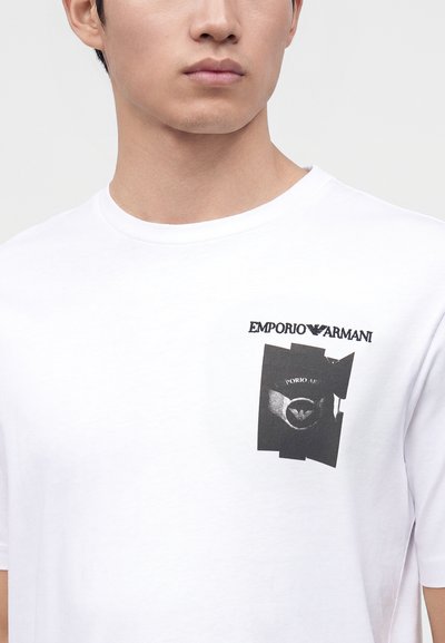 Άνδρας που φοράει λευκό T-shirt Emporio Armani με μαύρο γραφικό λογότυπο στο αριστερό στήθος, σε απλό ανοιχτό γκρι φόντο.