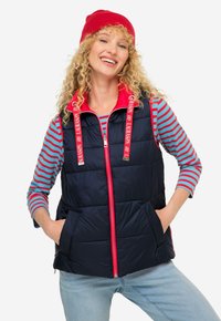 Marinblå pufferväst med röda accentuerande detaljer, med hög krage, dragkedjestängning och två framfickor. Bärs över en randig långärmad tröja.