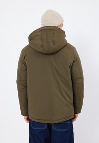 Chaqueta acolchada de color verde oliva con capucha, que presenta una textura suave y detalles de costuras. Llevada sobre pantalones oscuros de mezclilla y un gorro de punto ligero.