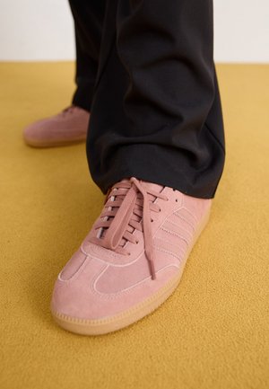 Personne portant des baskets en daim rose avec des semelles en gomme et un pantalon noir, debout sur un sol recouvert de moquette jaune.