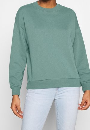 Felpa verde in un tessuto morbido con scollo rotondo, spalle ribassate e polsini a costine. Abbinata a jeans azzurri per un look rilassato.