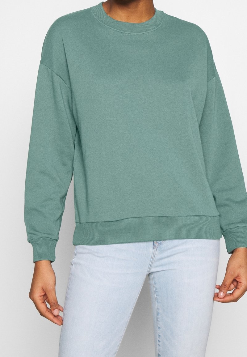 Sweatshirt vert en tissu doux avec un col rond, des épaules tombantes et des poignets côtelés. Associé à un jean bleu clair pour un look décontracté.